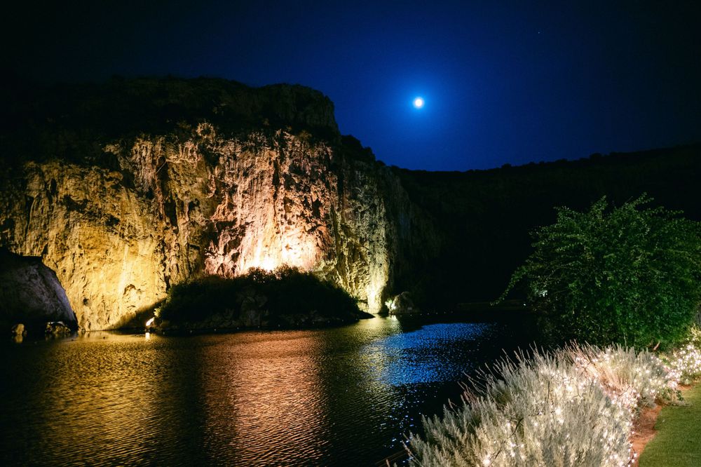 Dramatic nighttime landscape with illuminated cliffs and full moon reflecting on calm waters.
