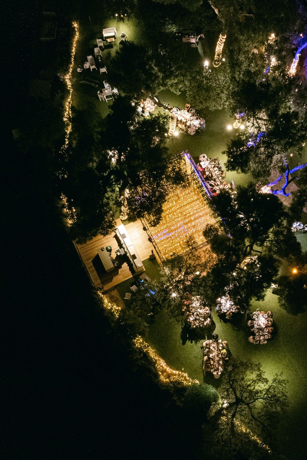 Aerial view of an outdoor wedding reception with twinkling lights and tables arranged under trees.