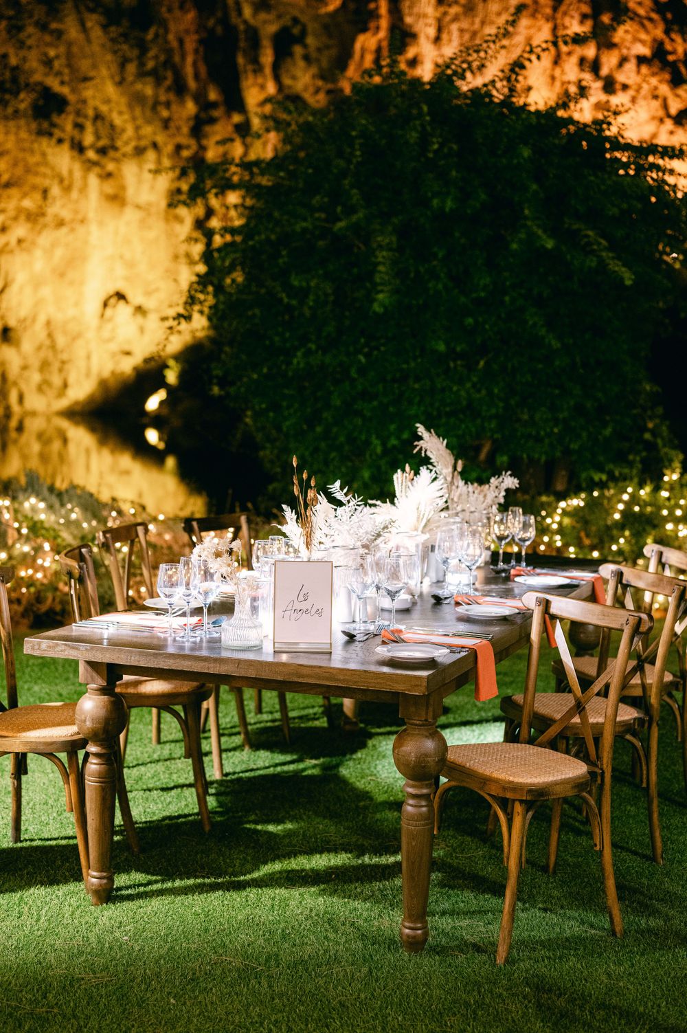Rustic wedding table setting with wooden chairs and elegant place settings against illuminated rock wall.