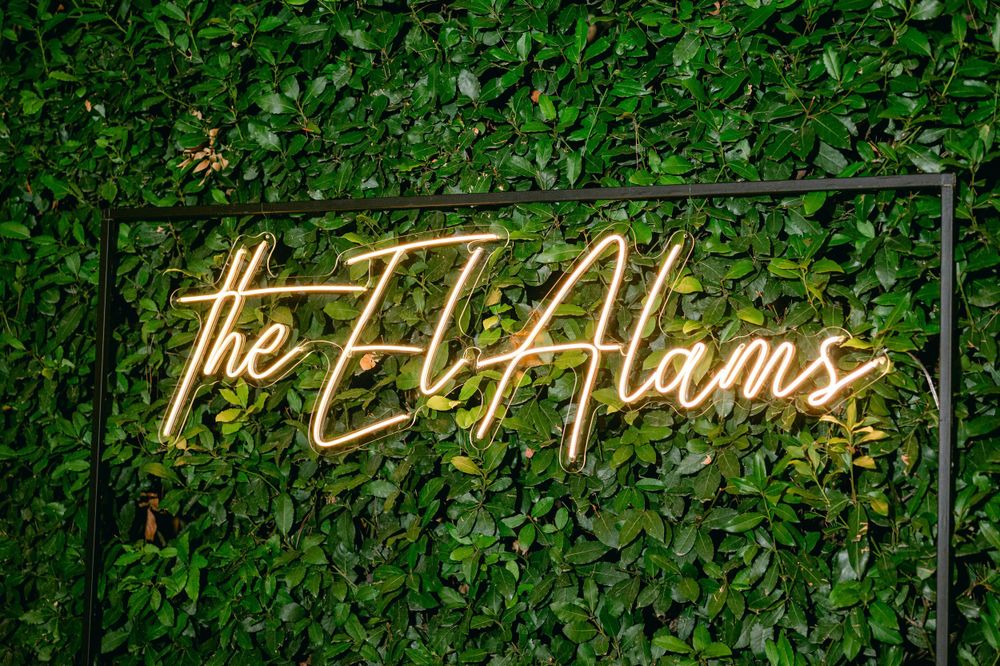 Neon sign reading 'The El Alams' mounted on green foliage wall.