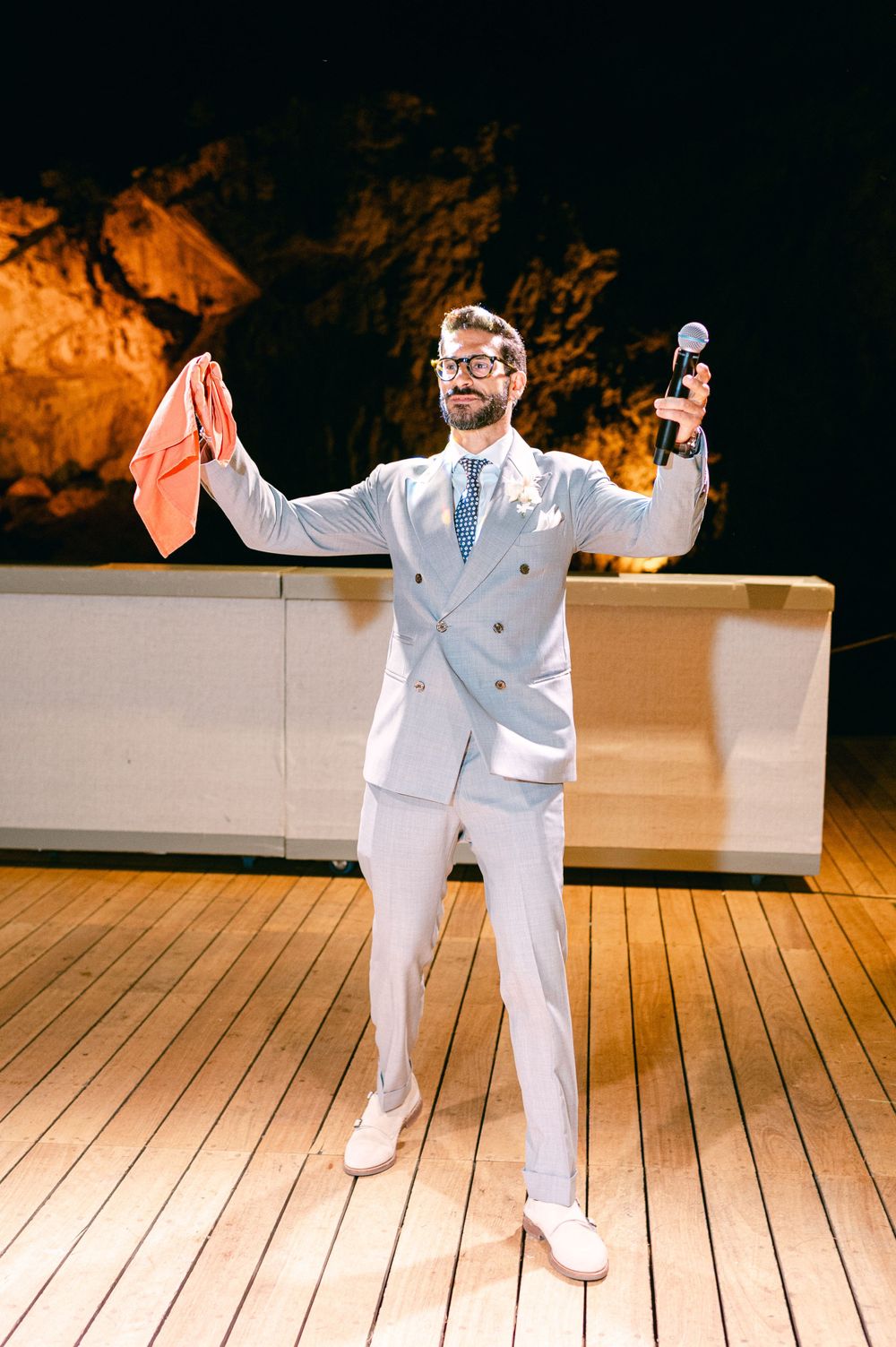 Wedding guest in white suit holding napkin and microphone against lit rocky backdrop.