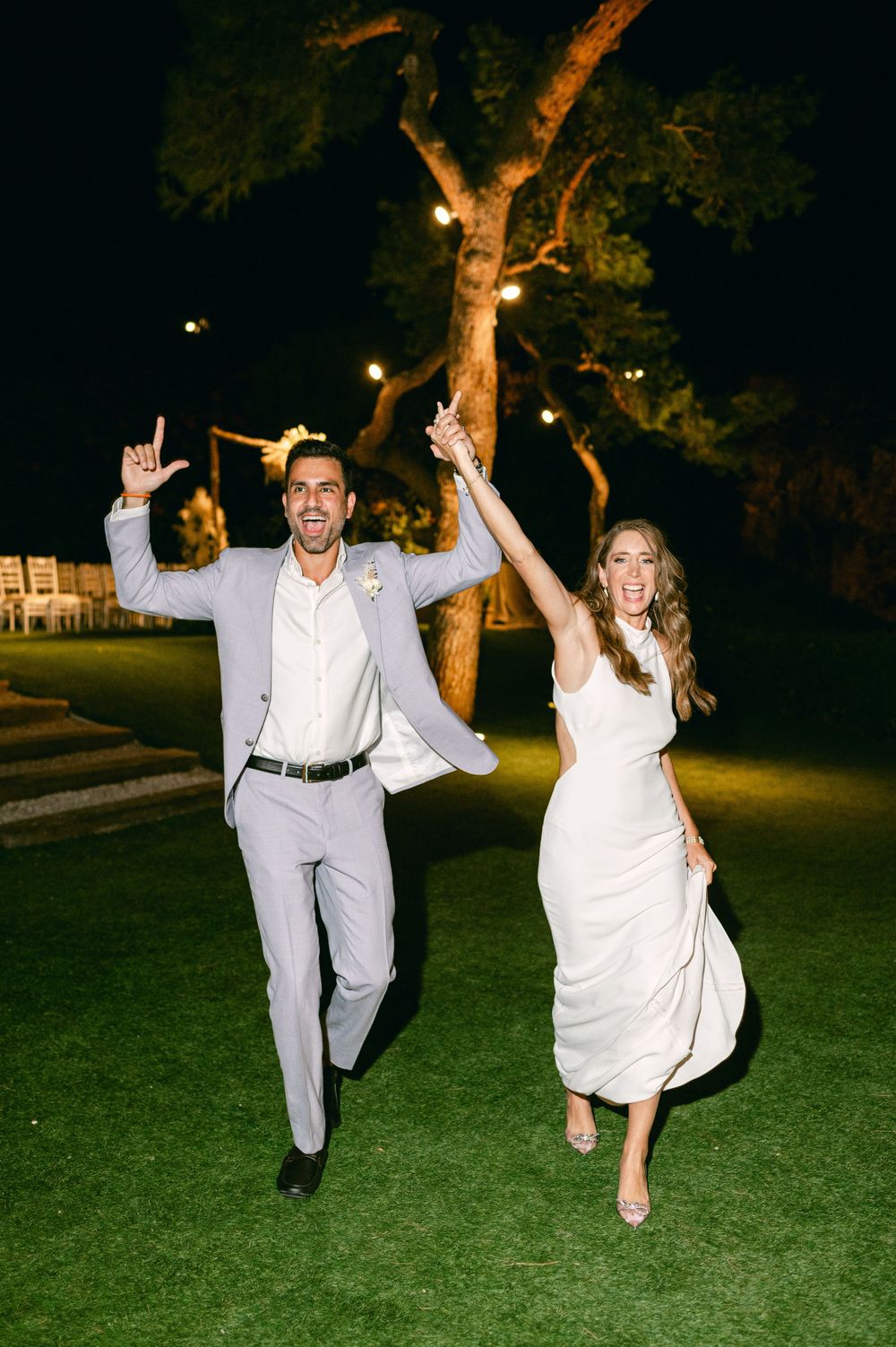 Joyful couple celebrating on grass under evening lights at outdoor wedding reception.