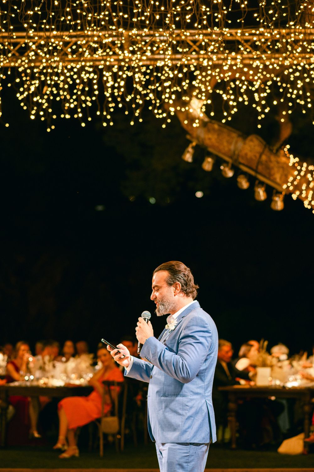 Speaker in light blue suit addresses guests under warm glowing string lights at outdoor venue.