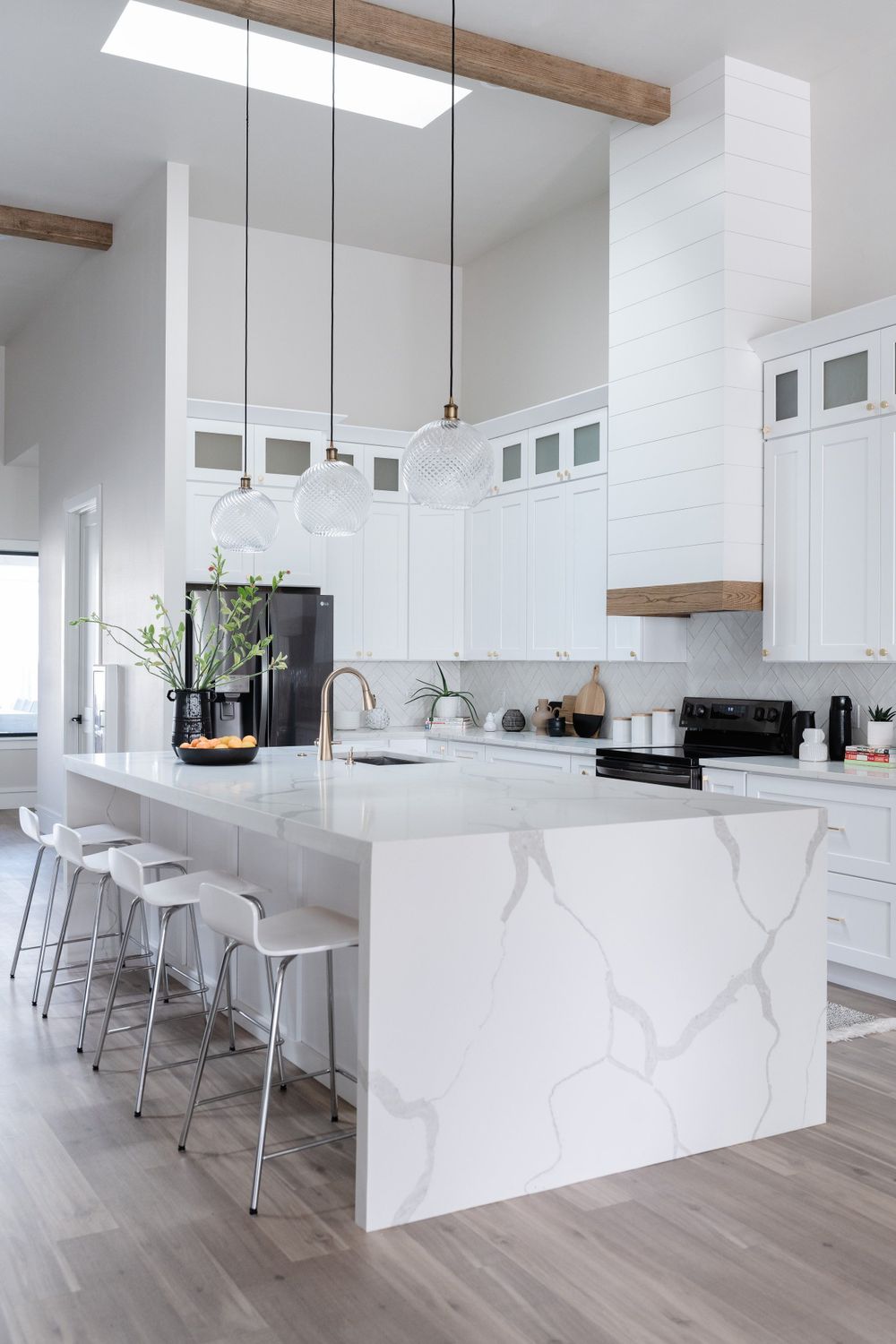 Modern white kitchen with marble waterfall island, wooden beams, and pendant lights.