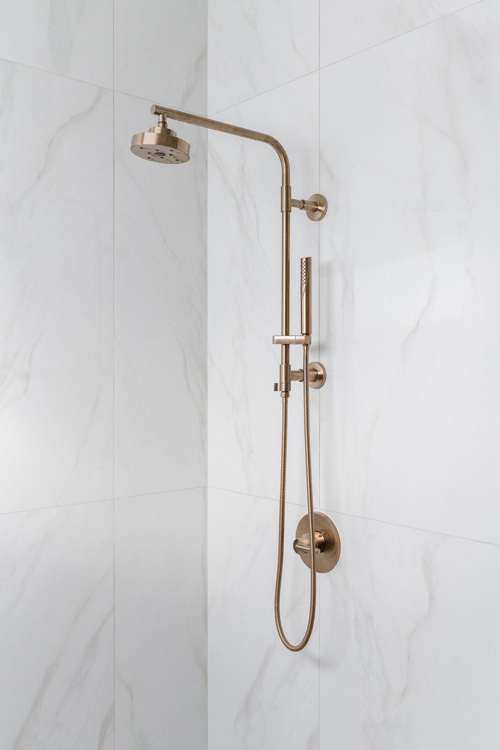 Luxury brass shower fixture with rainhead and handheld sprayer mounted on white marble wall.