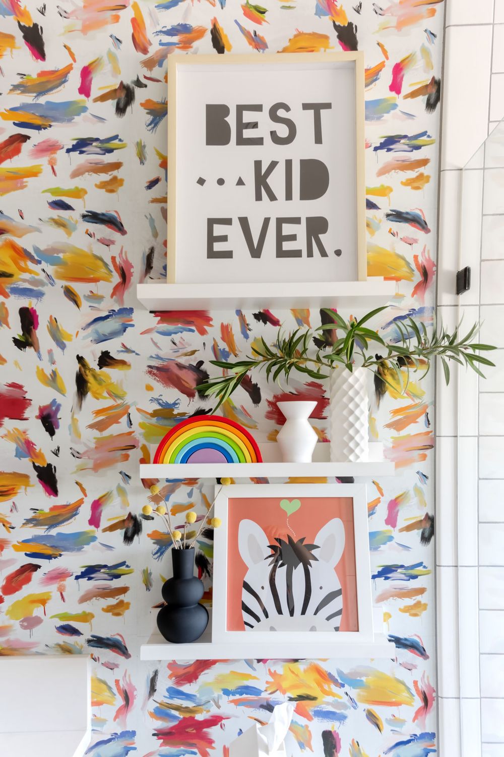 Colorful abstract wallpaper with 'Best Kid Ever' sign and rainbow decor on white floating shelf.