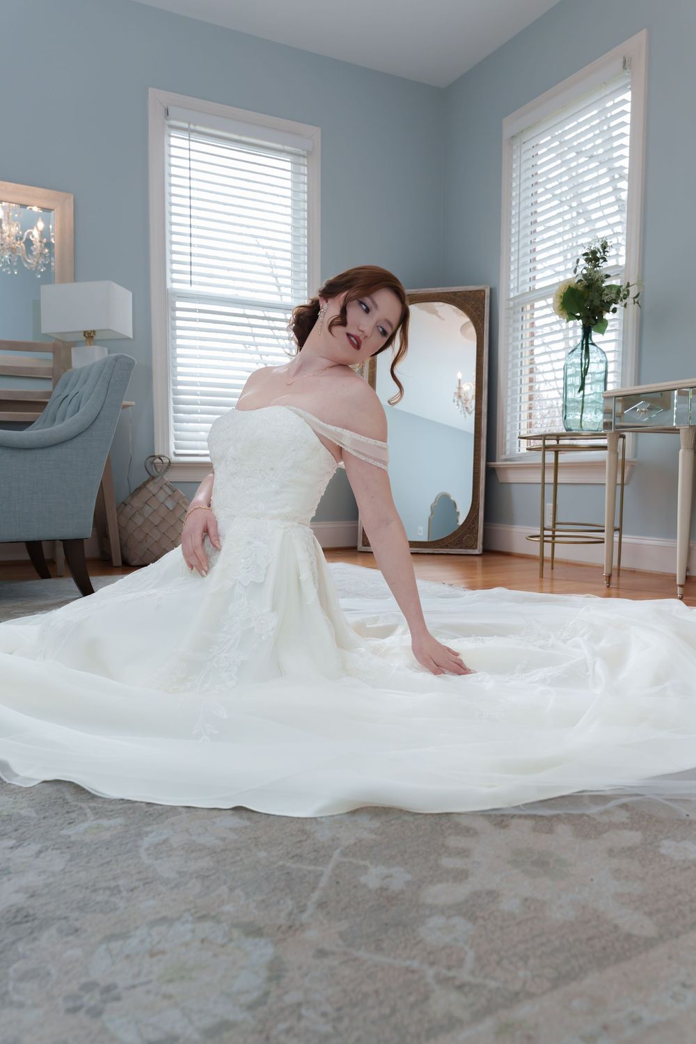 A bride in a flowing white strapless gown poses gracefully on the floor in a blue-walled room with natural lighting.