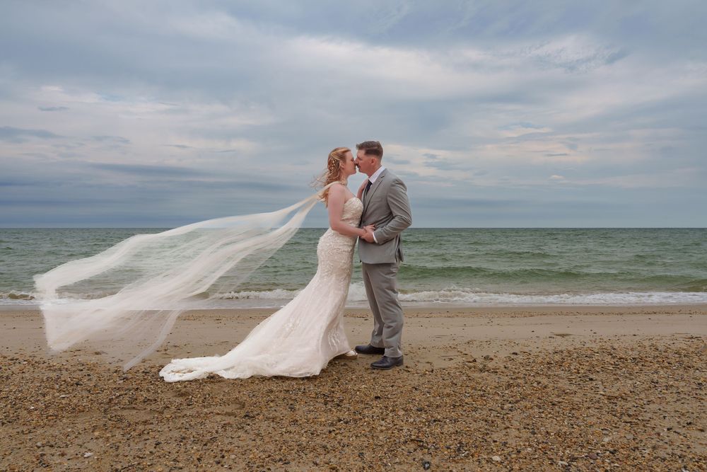 Bride and groom share a romantic kiss on a sandy beach, with the bride's veil flowing in the wind against a cloudy sky.