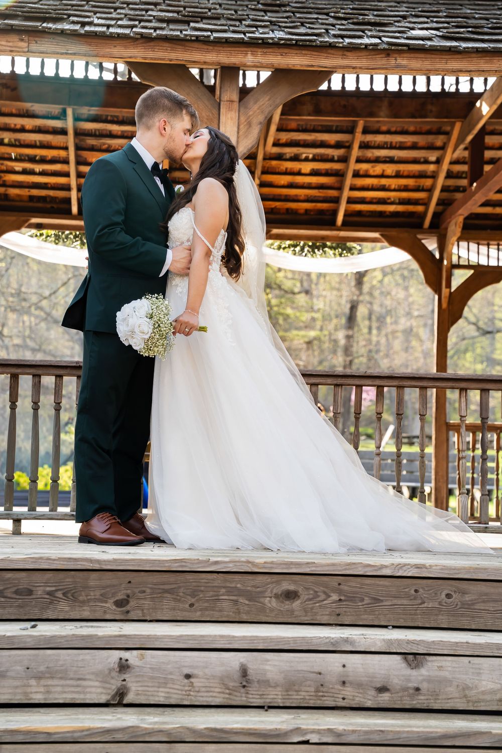 Wedding couple sitting on rustic wooden benches under a covered pavilion with white floral bouquet.