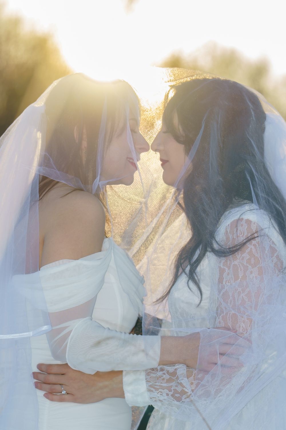Two brides in white wedding attire share an intimate moment in golden sunlight during their outdoor ceremony.