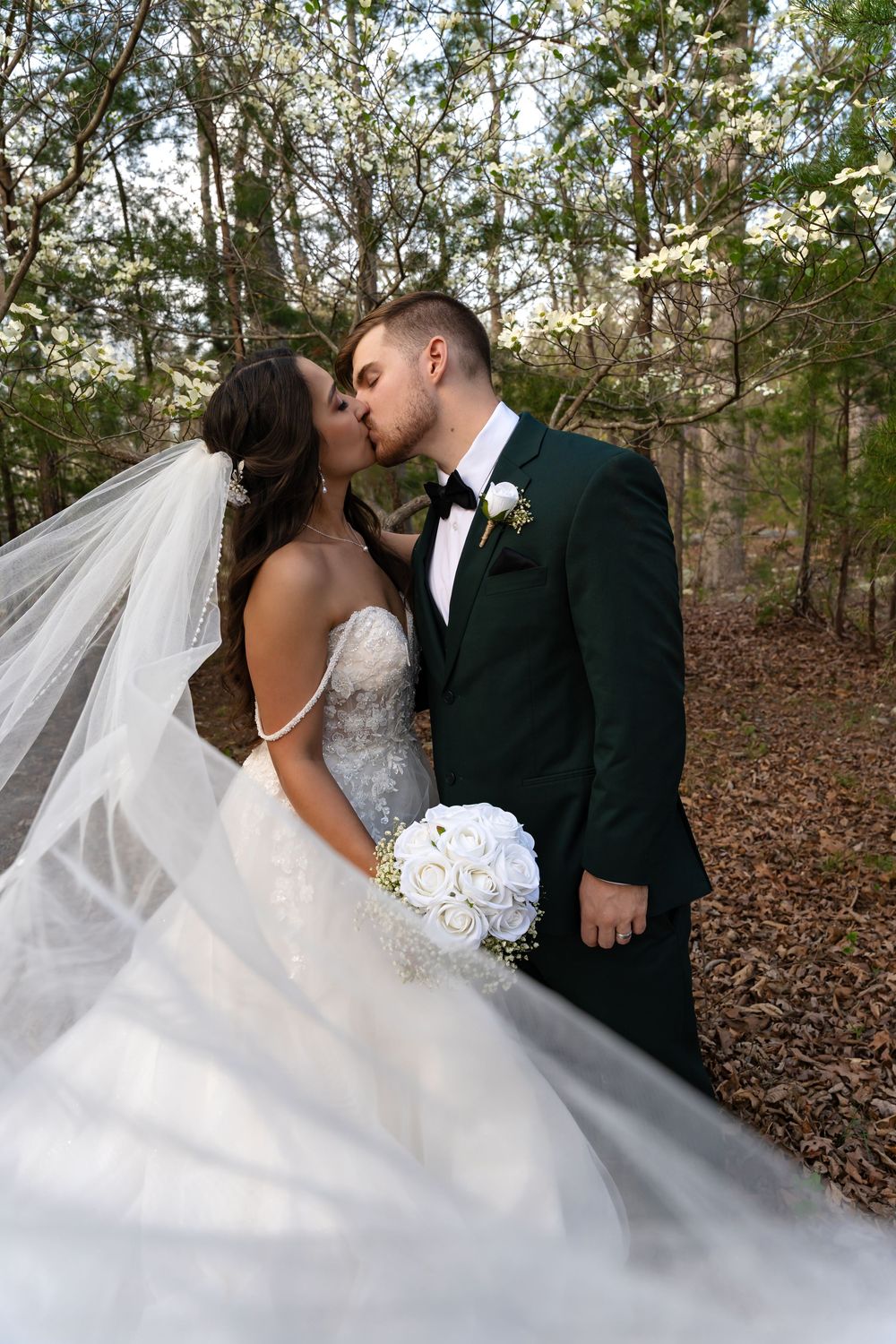 A wedding couple embraces in a romantic moment surrounded by flowering trees.