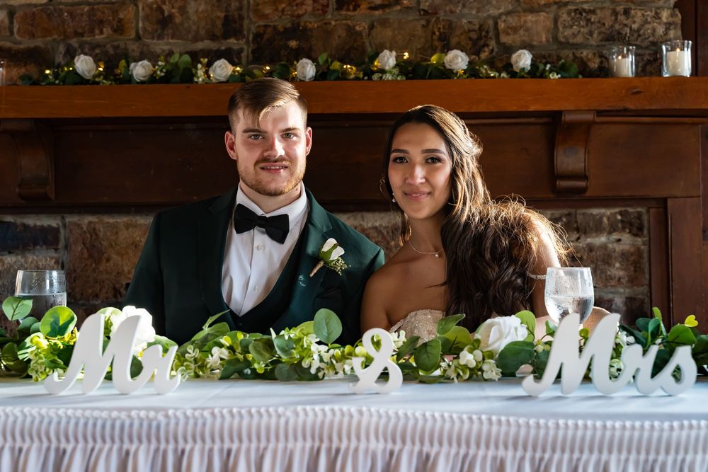 A newlywed couple sits at a decorated reception table with 'Mr & Mrs' signage.