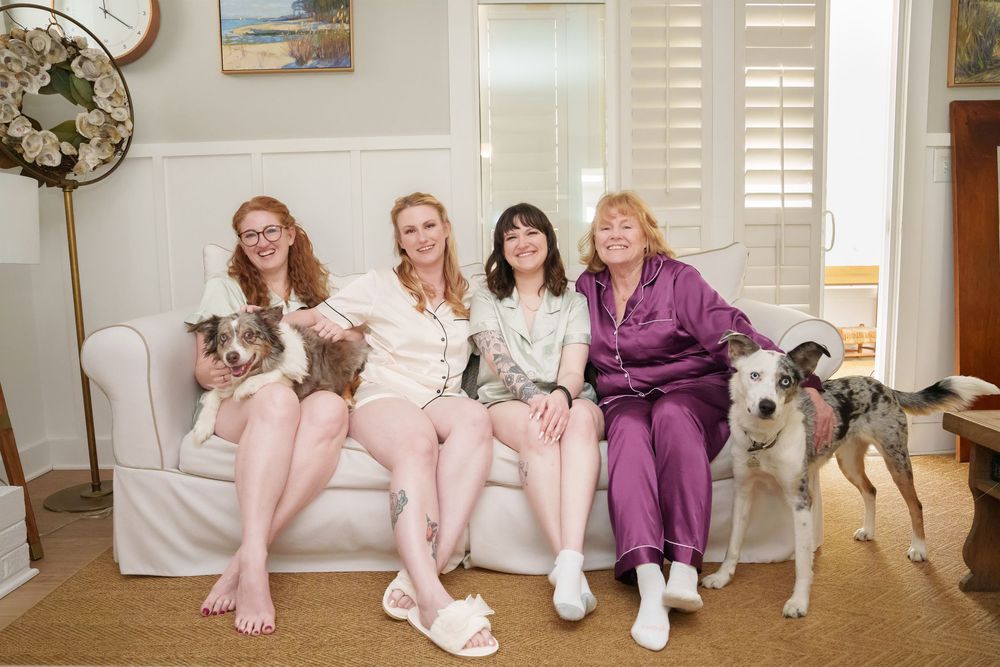 Four women in pajamas sitting on a white sofa with two greyhound dogs in a cozy living room.
