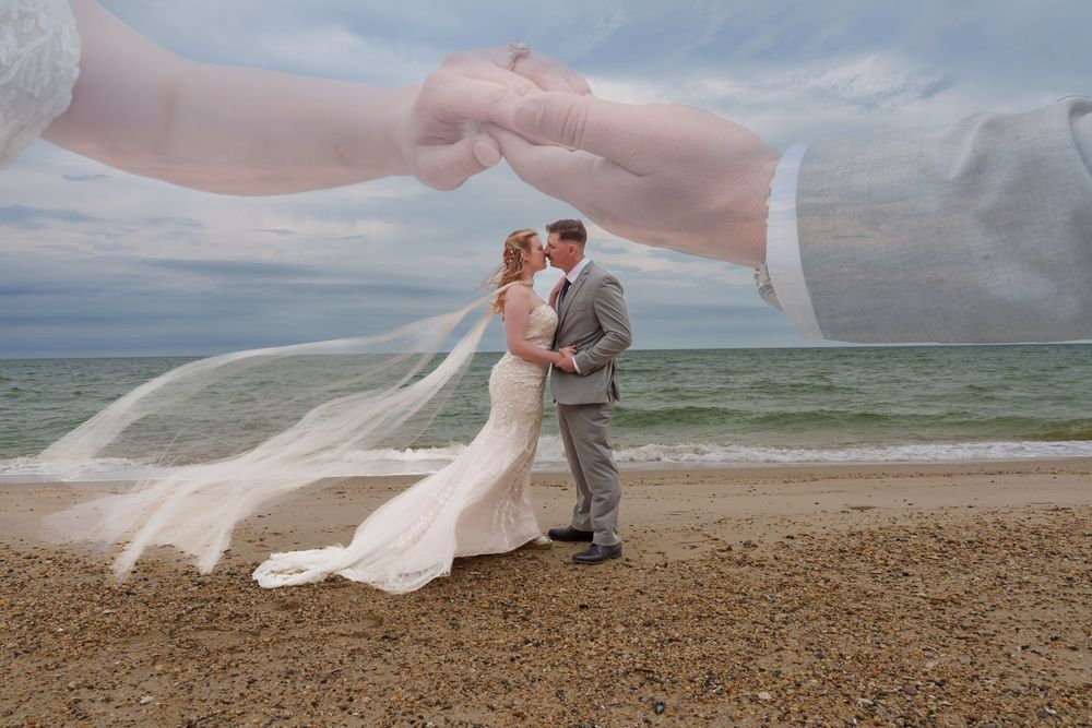 Bride and groom share a romantic kiss on a beach, her veil flowing in the wind, with ocean waves in the background.
