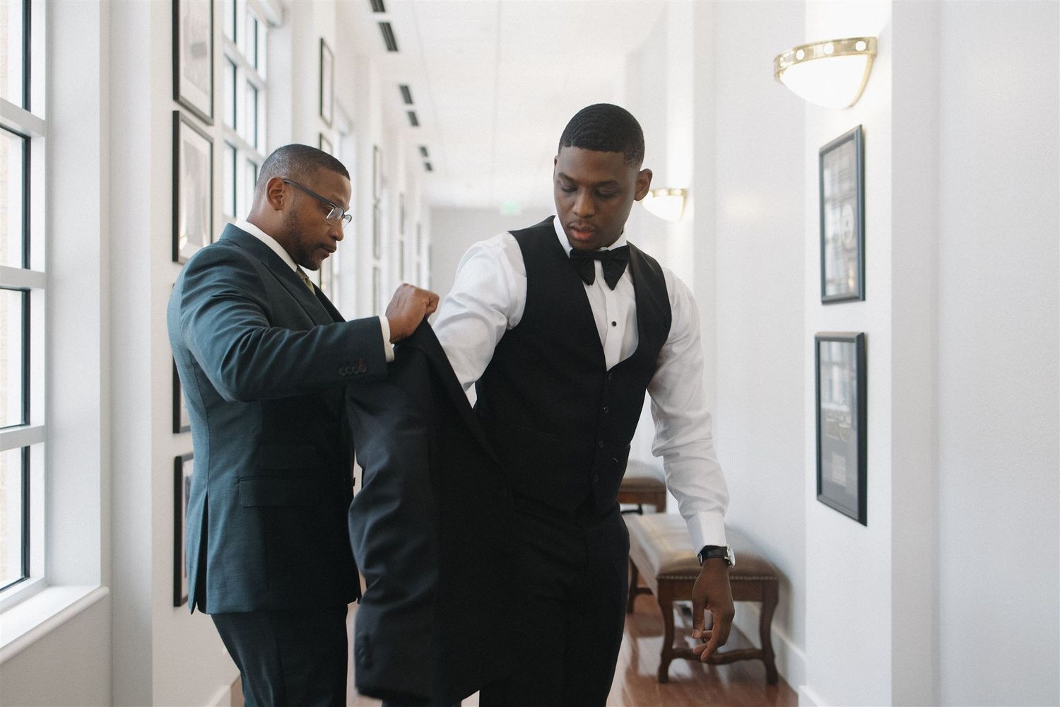 Two people in formal attire help adjust a black vest in a bright white hallway with framed artwork.