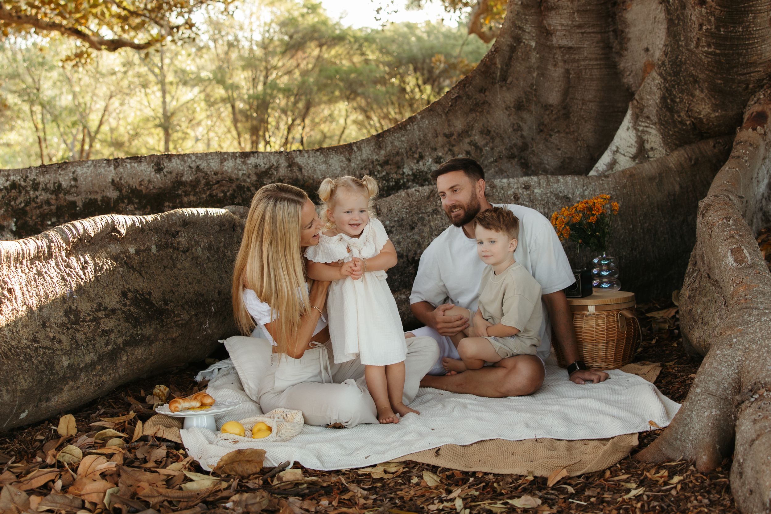 Two children sit together on a blanket near large tree roots with a vase of flowers on a small table beside them.