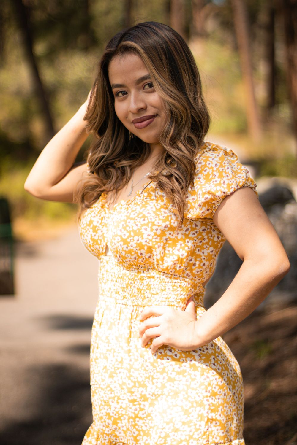 Woman in yellow floral dress poses outdoors in natural garden setting.