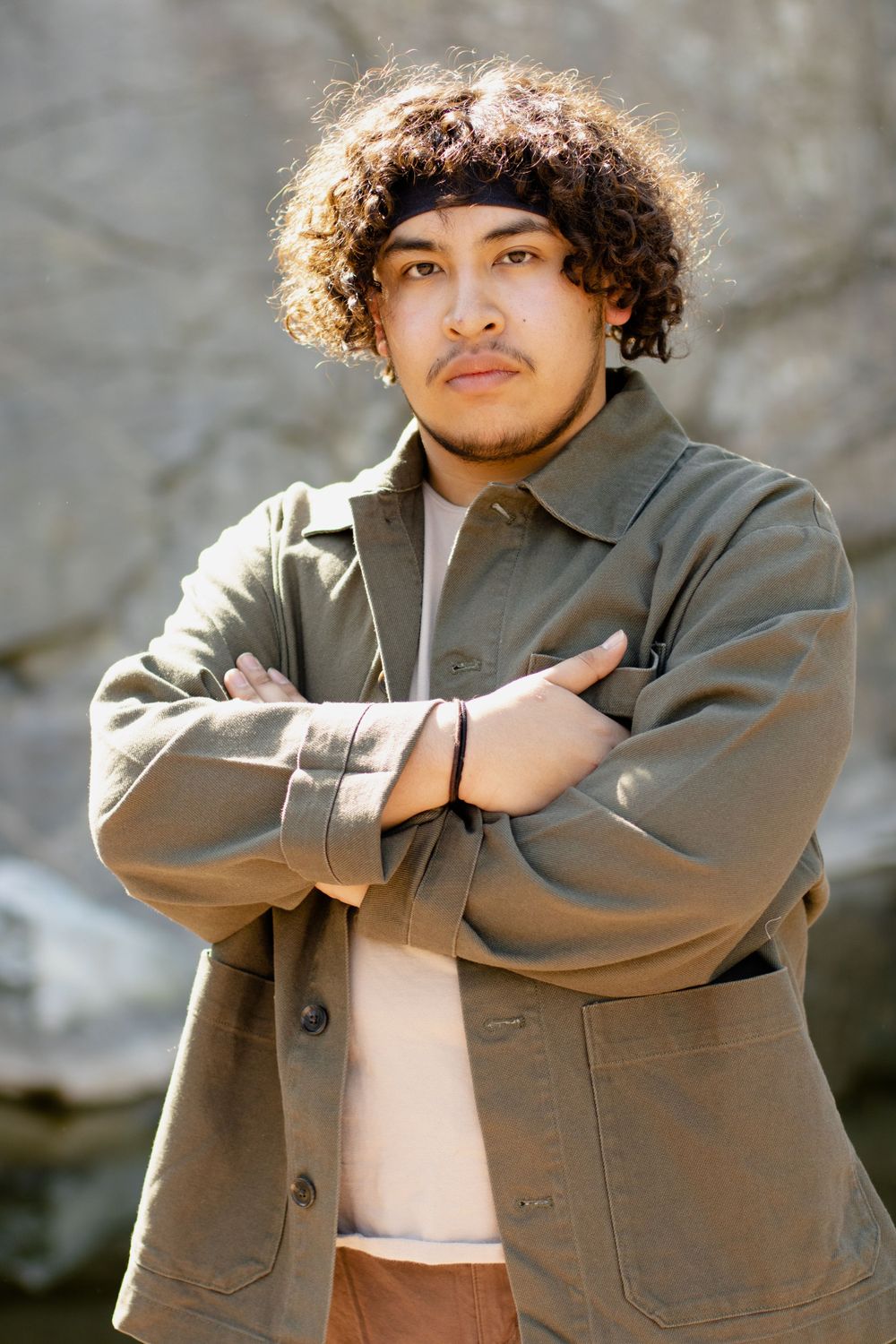Person wearing brown jacket poses with arms crossed against stone wall background.