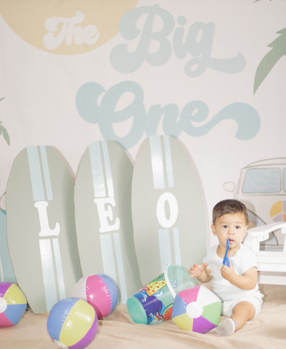 Baby sitting by surfboard wall decor with colorful beach balls and vintage-themed party decorations.