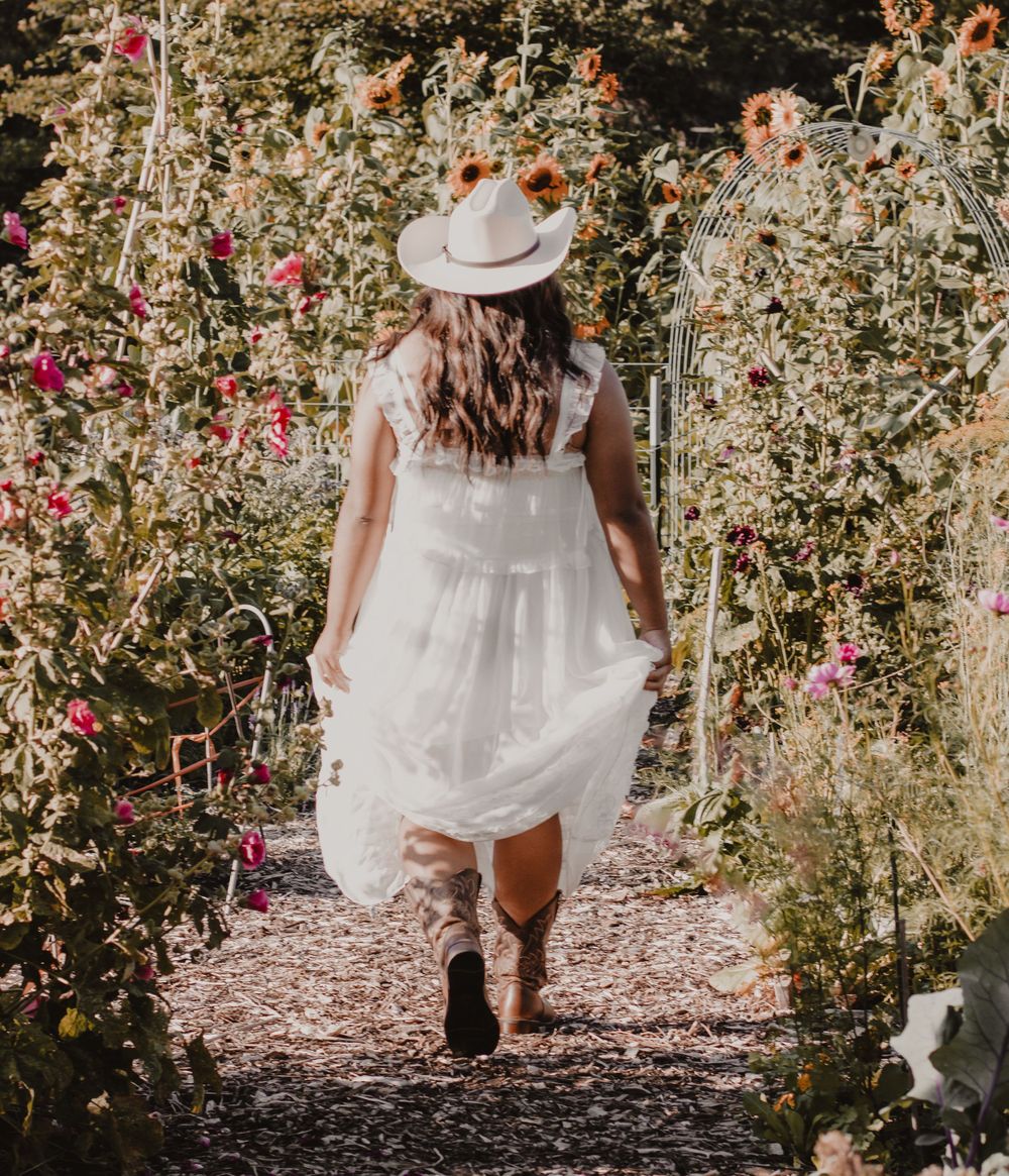 Woman in white dress and hat walks through garden path lined with pink flowers in dreamy summer setting.