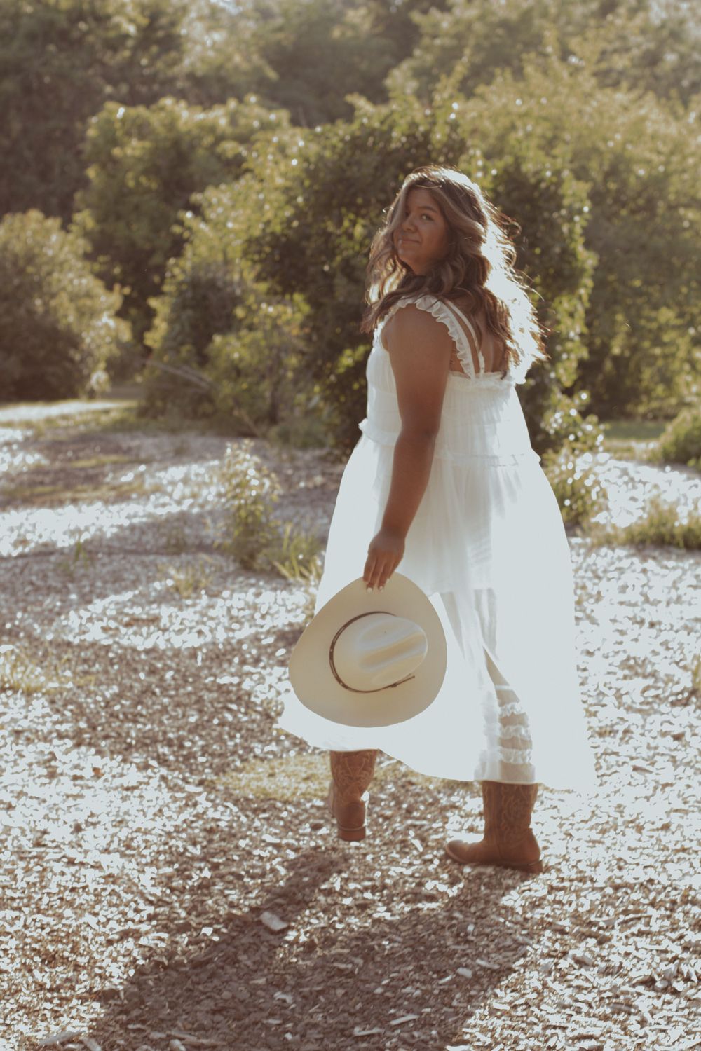 Woman in flowing white dress holds straw hat while standing on sunlit dirt path.