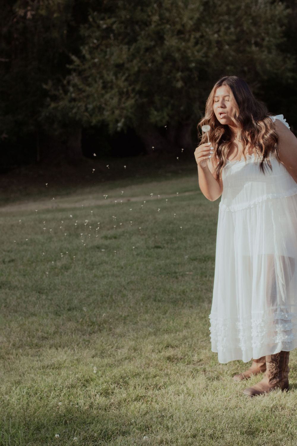 Woman in white dress holds flowers in sunny meadow wearing western boots.