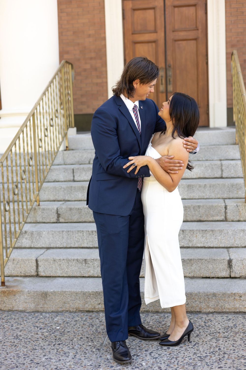 A couple embraces on stone steps outside a building with wooden doors and brick walls.