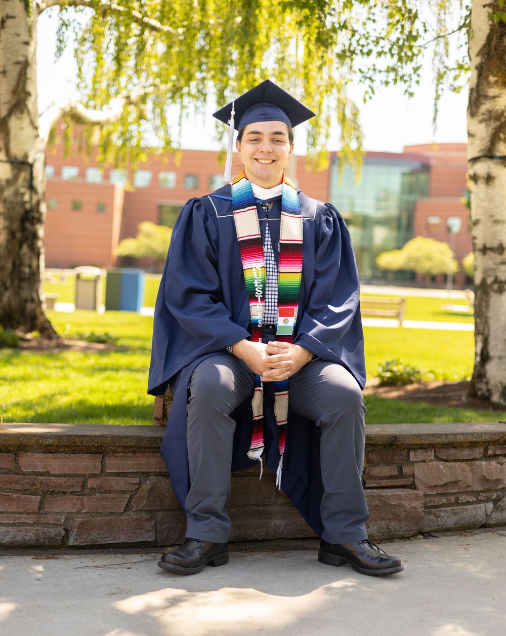Graduate in cap and gown with colorful stole sits on stone wall with campus buildings in background.