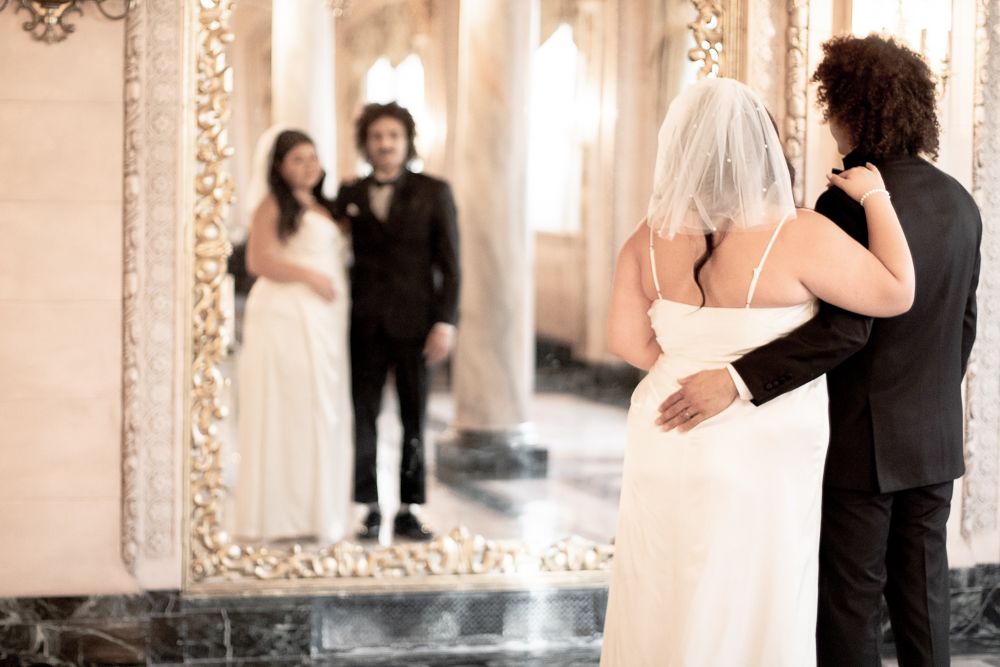 Couple in wedding attire looks at their reflection in ornate mirror.