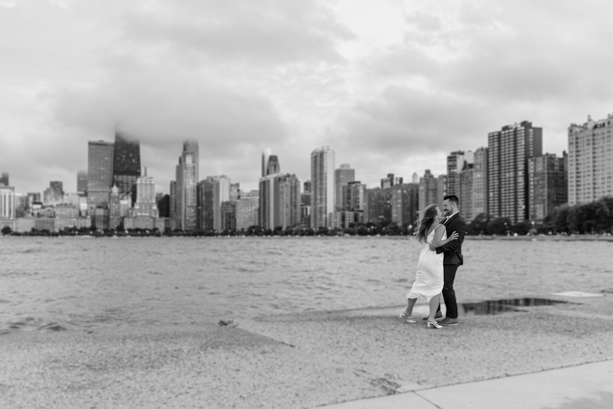 A couple embraces on a beach with the Chicago skyline silhouetted in the background.