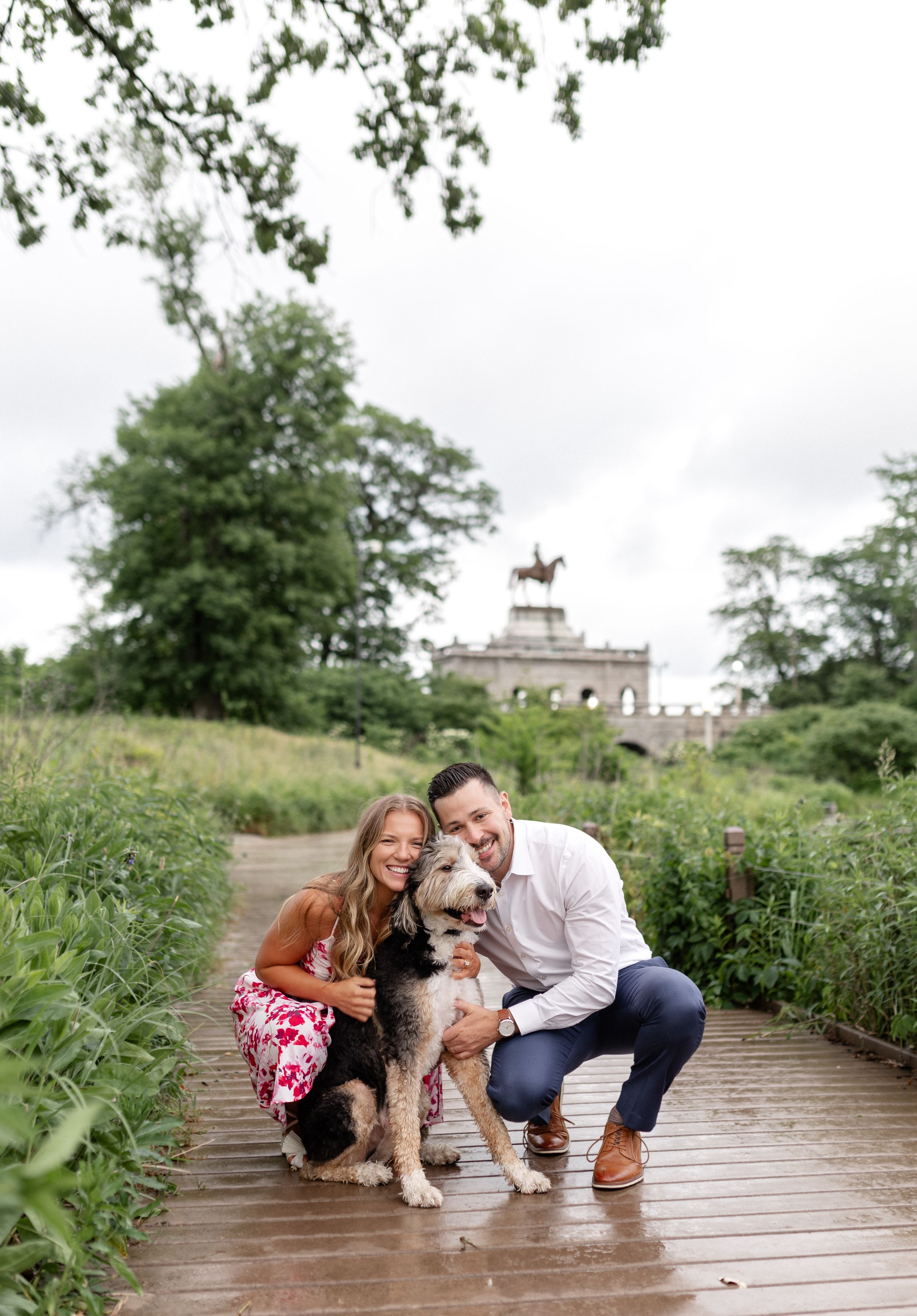 A couple poses with their dog on a wooden boardwalk with a historic building visible in the background.