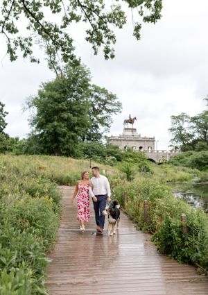 A couple walks their dog along a scenic wooden pathway with a landmark building in the distance.