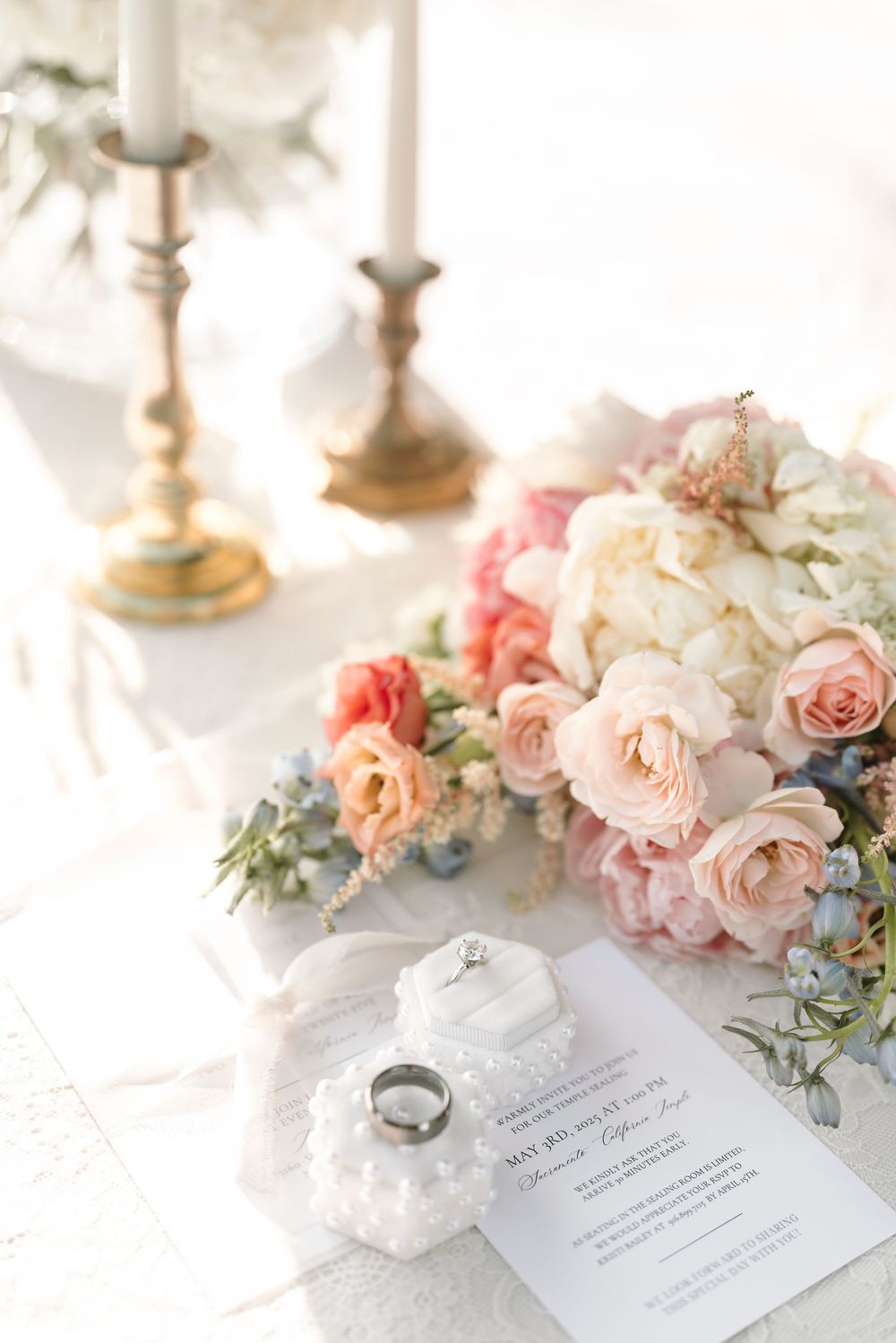 Elegant wedding details featuring blush roses, wedding rings and invitation on white linen with soft natural lighting.