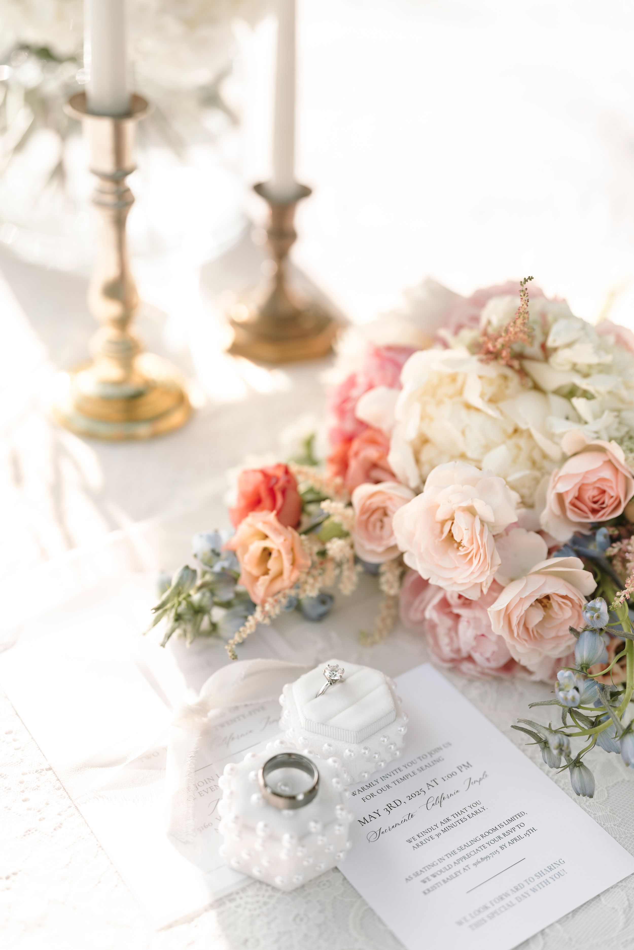 Elegant wedding details featuring blush roses, wedding rings and invitation on white linen with soft natural lighting.