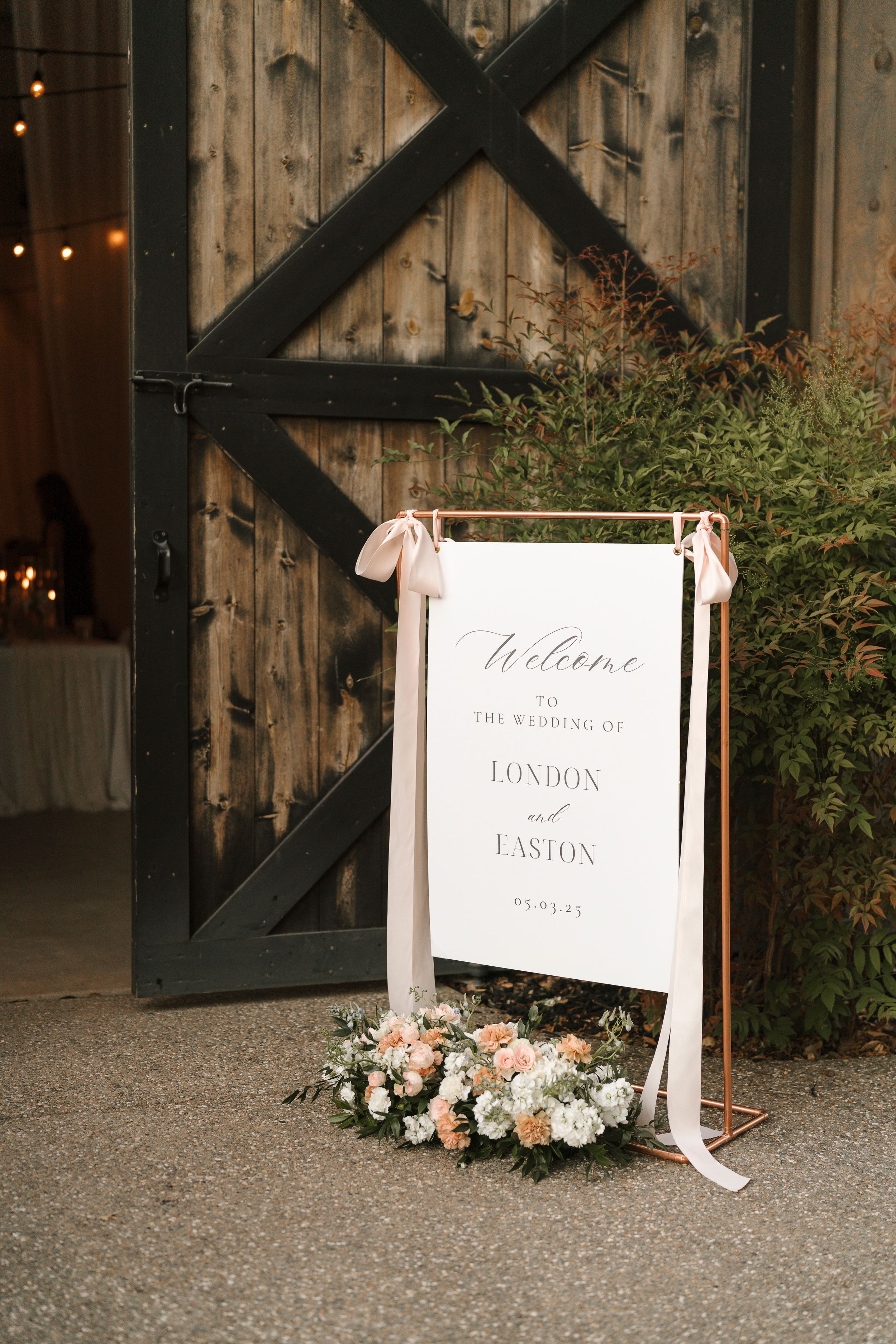 Elegant wedding welcome sign with floral arrangements and copper frame stands against rustic barn wood backdrop.