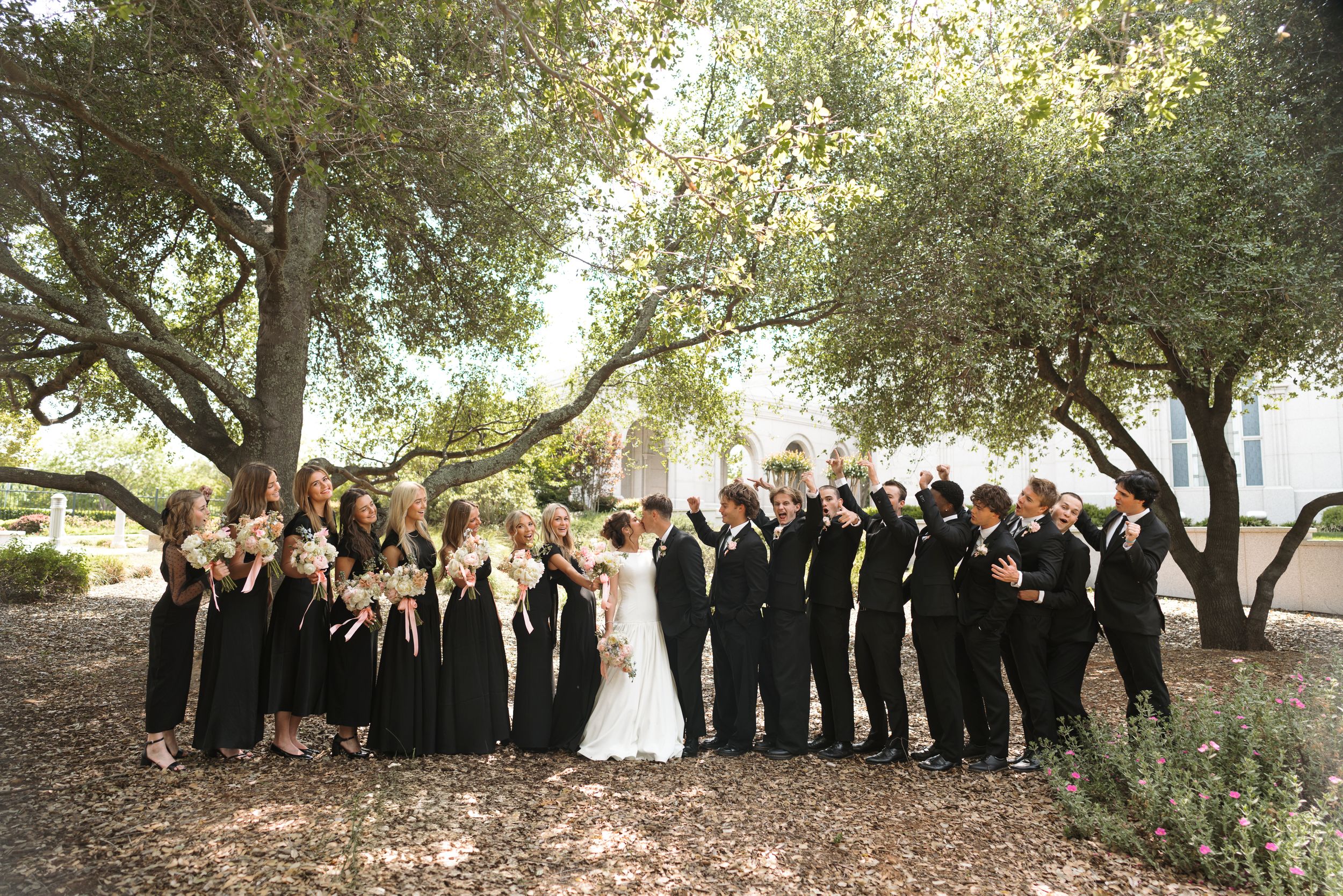 Wedding party members stand between trees at an outdoor celebration in formal attire.