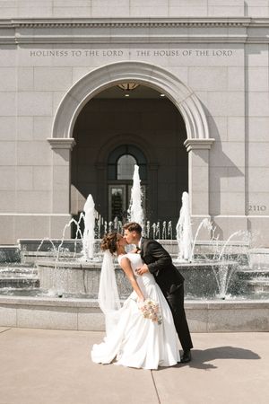 A romantic wedding couple poses together in front of a stone church building with fountains.