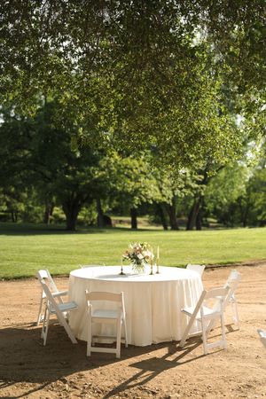 White folding chairs and round tables draped in white linens set up for an outdoor event under shady trees.