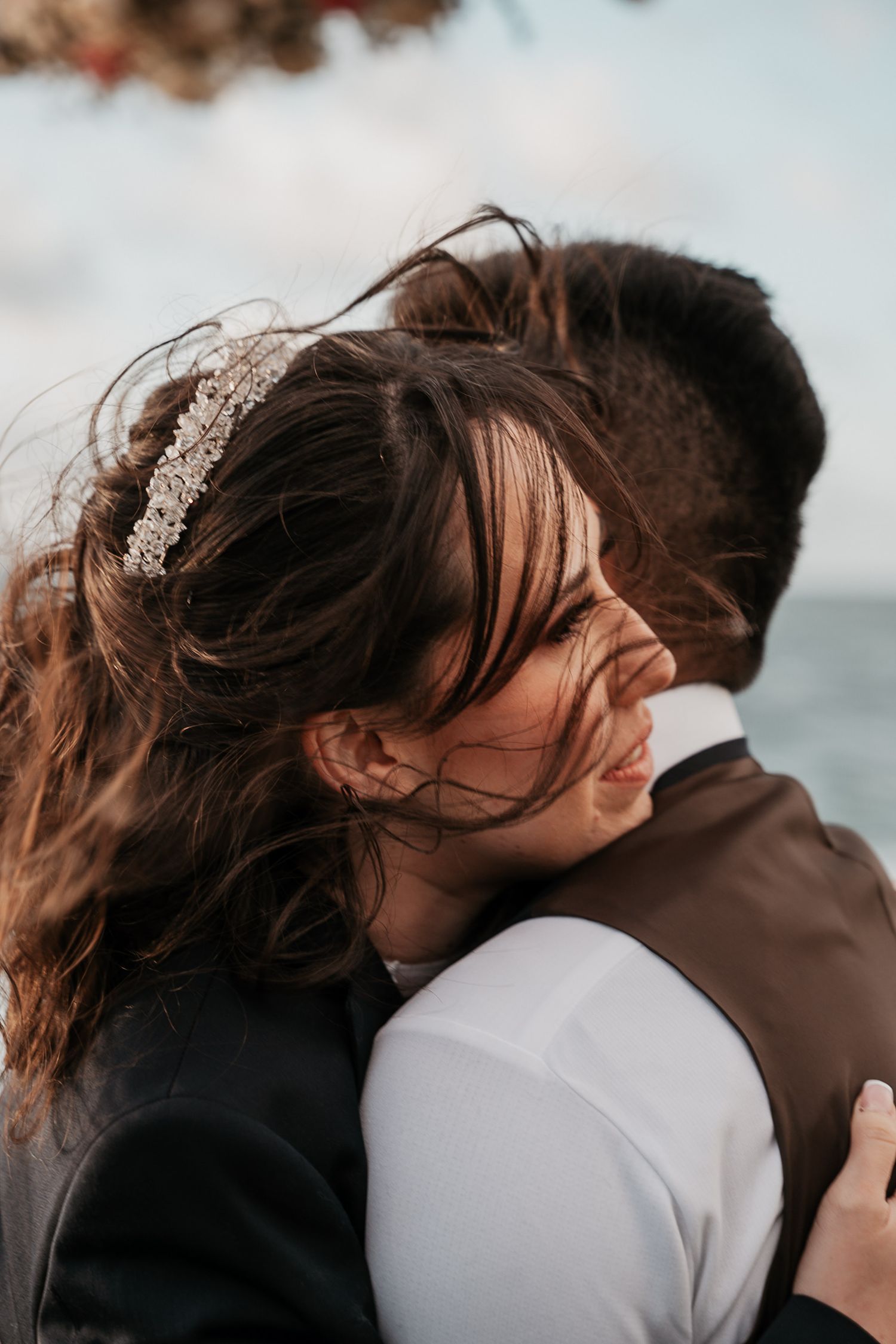 A romantic moment captured as a couple embraces near the ocean, with the bride wearing a delicate crystal headpiece.