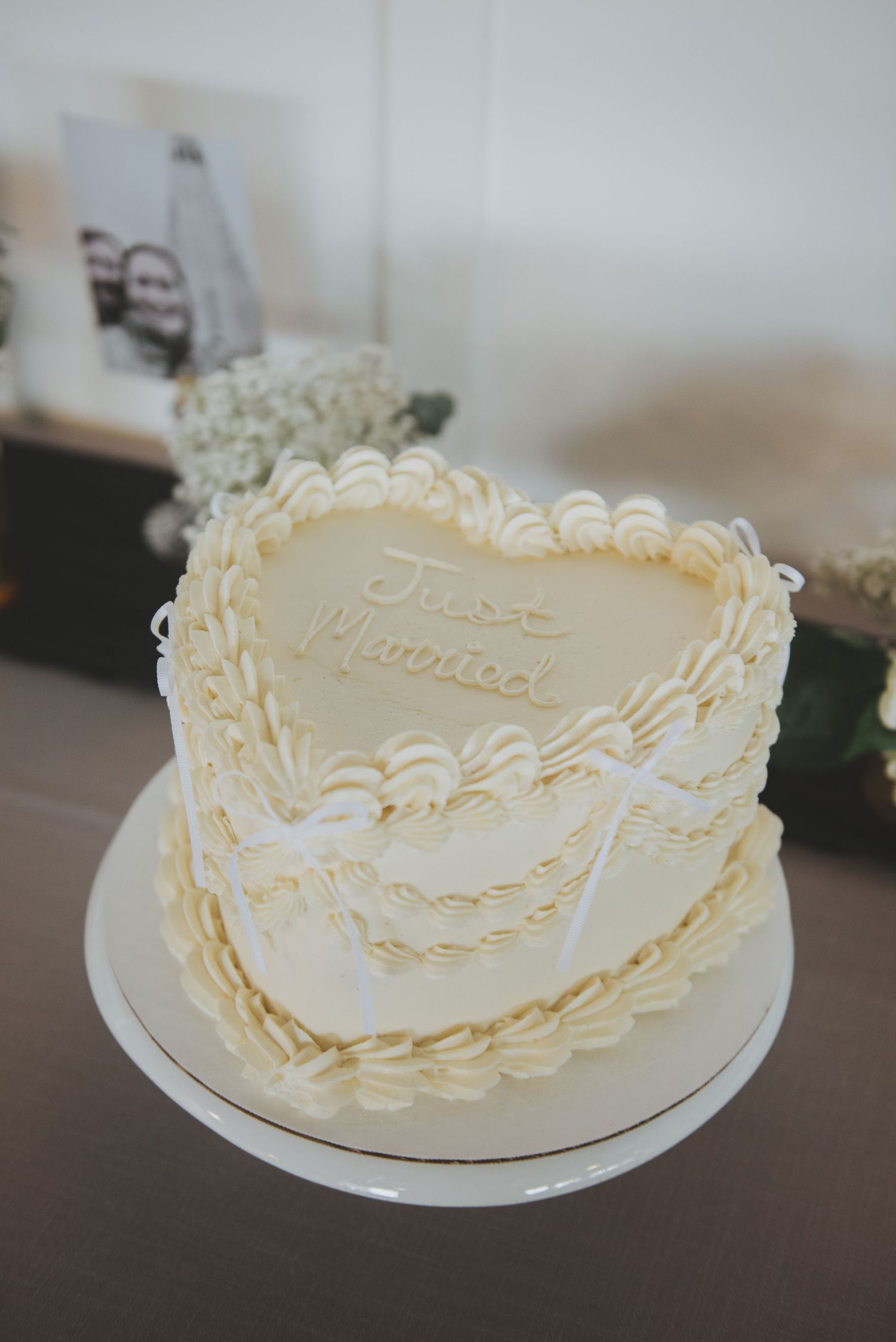 A heart-shaped white wedding cake with delicate piped lace details and elegant scalloped decorations sits on a simple cake stand.