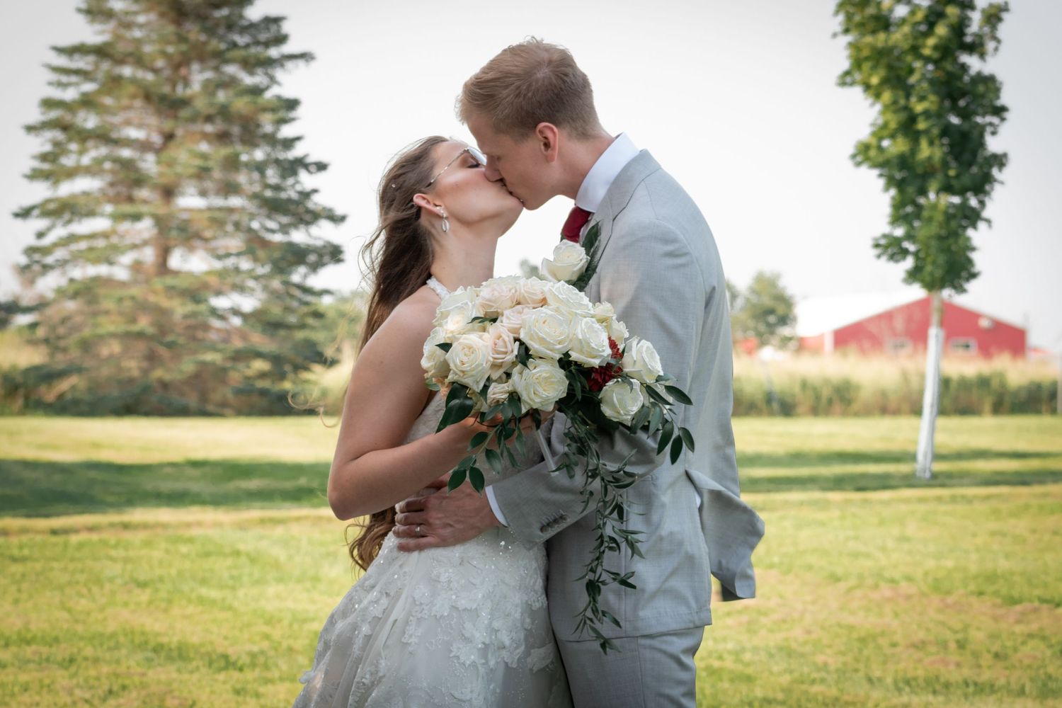 A bride and groom kiss in a field. She is holding her flowers. There is a red barn in the background
