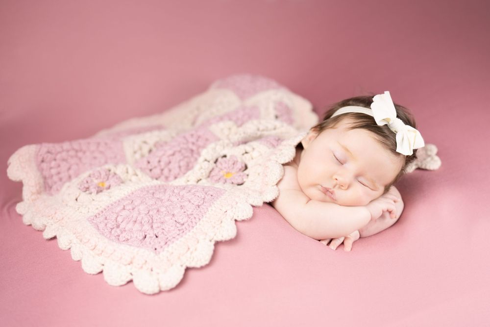 Newborn baby peacefully sleeping under a pink crocheted blanket with white bow headband.