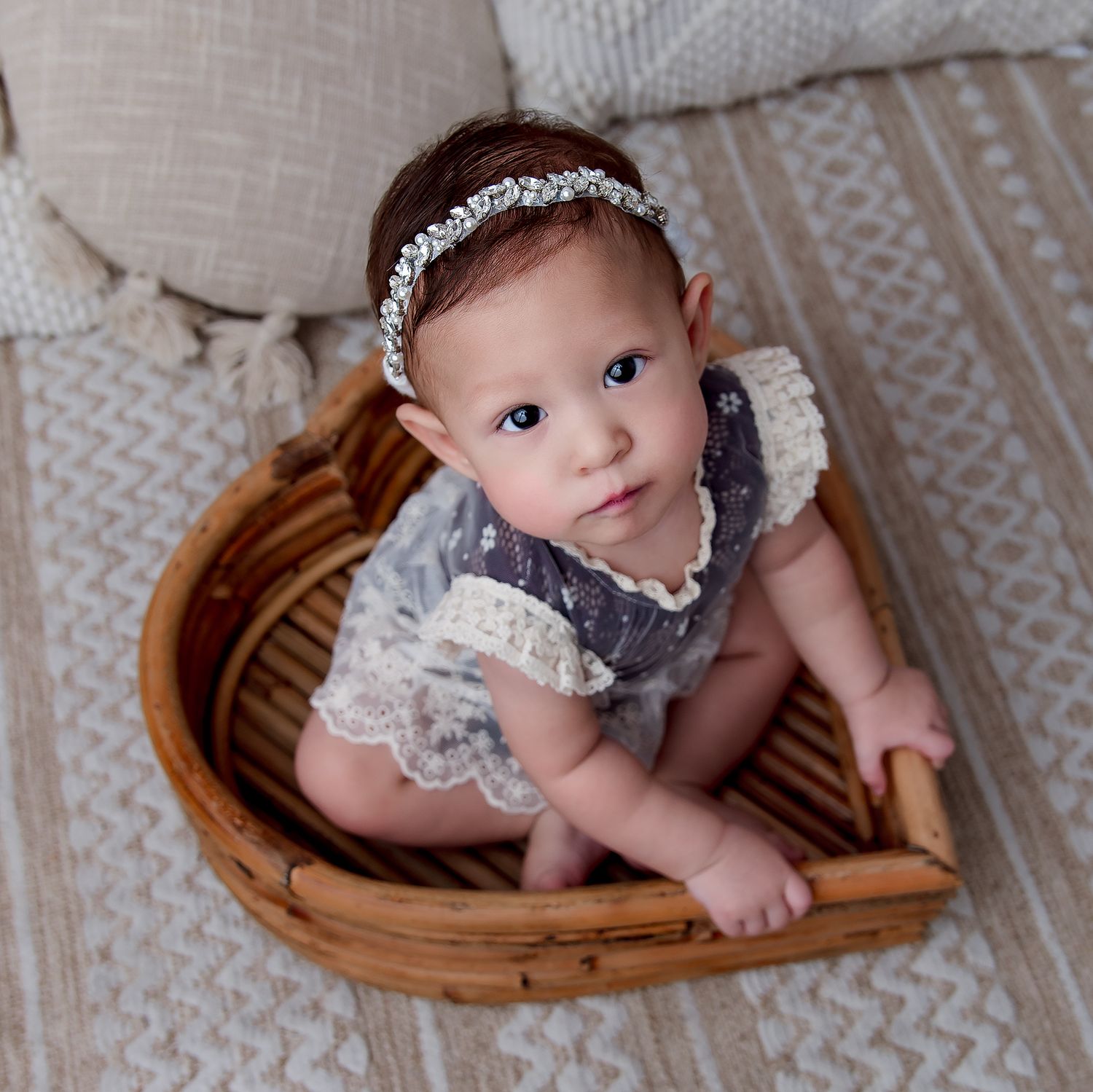 Baby in lace dress and crystal headband sitting in woven basket.