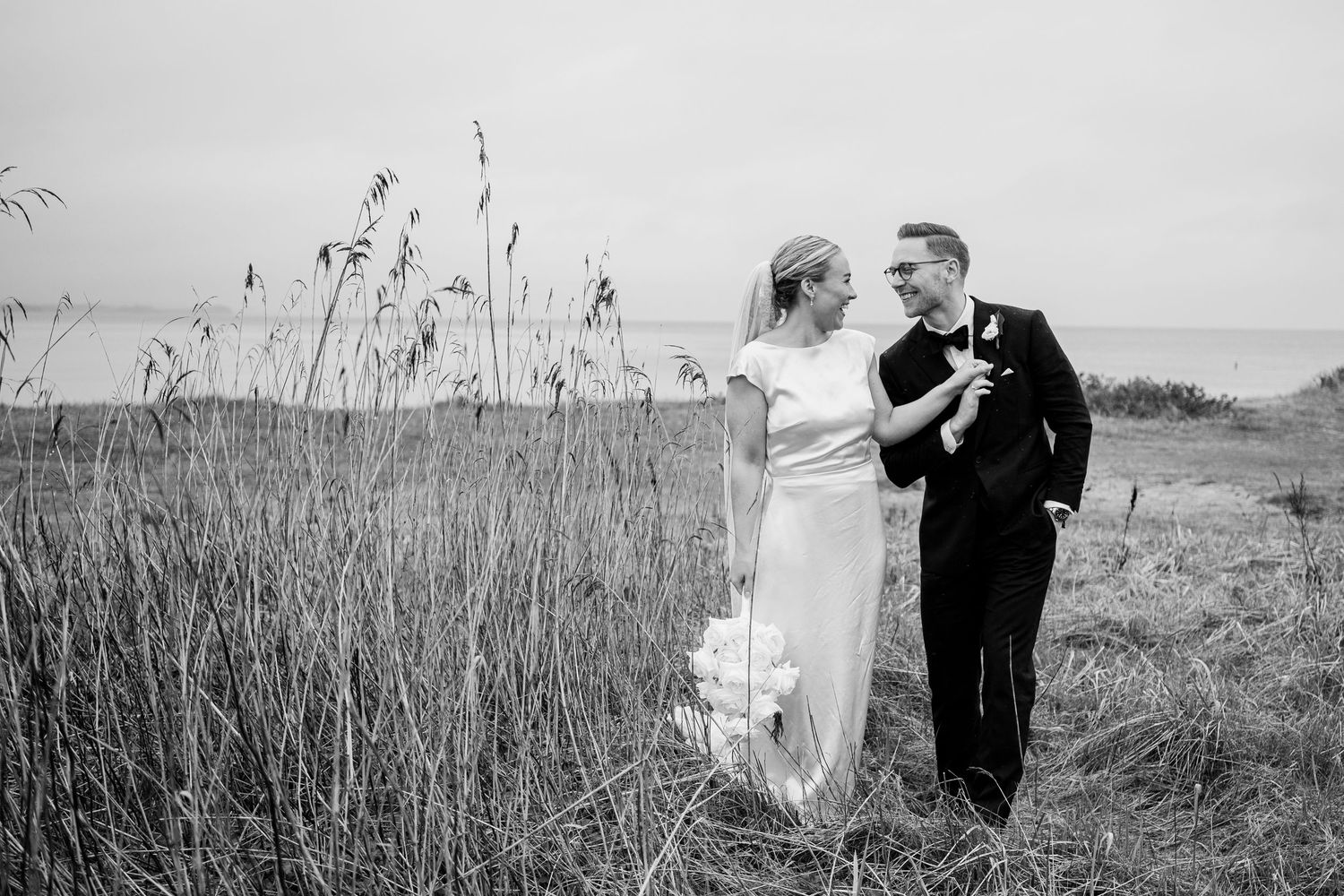 Newlyweds walking hand in hand through a sunlit forest wearing elegant wedding attire.
