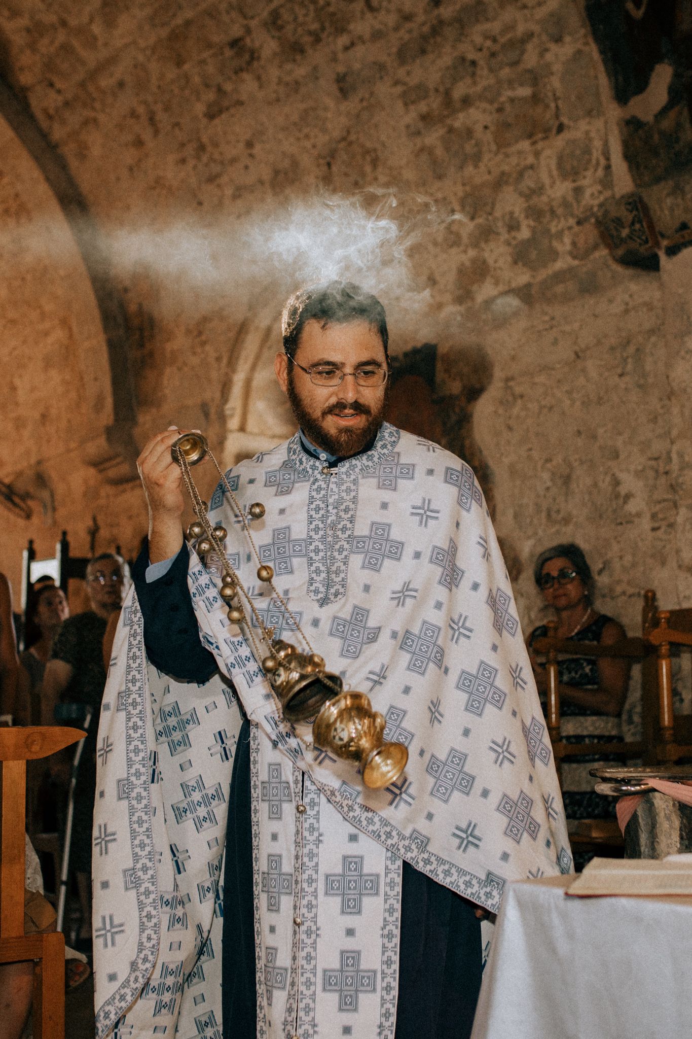 Orthodox priest in white embroidered robes swings a ceremonial censer during religious service in stone church interior. Cyprus