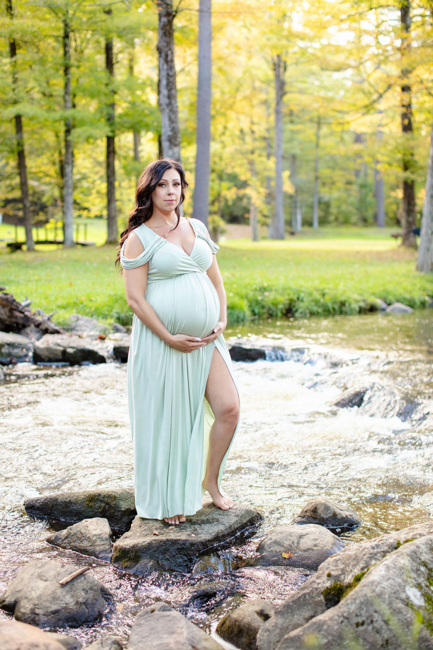 Maternity photo at waterfall showing woman in flowing mint dress amid autumn trees.
