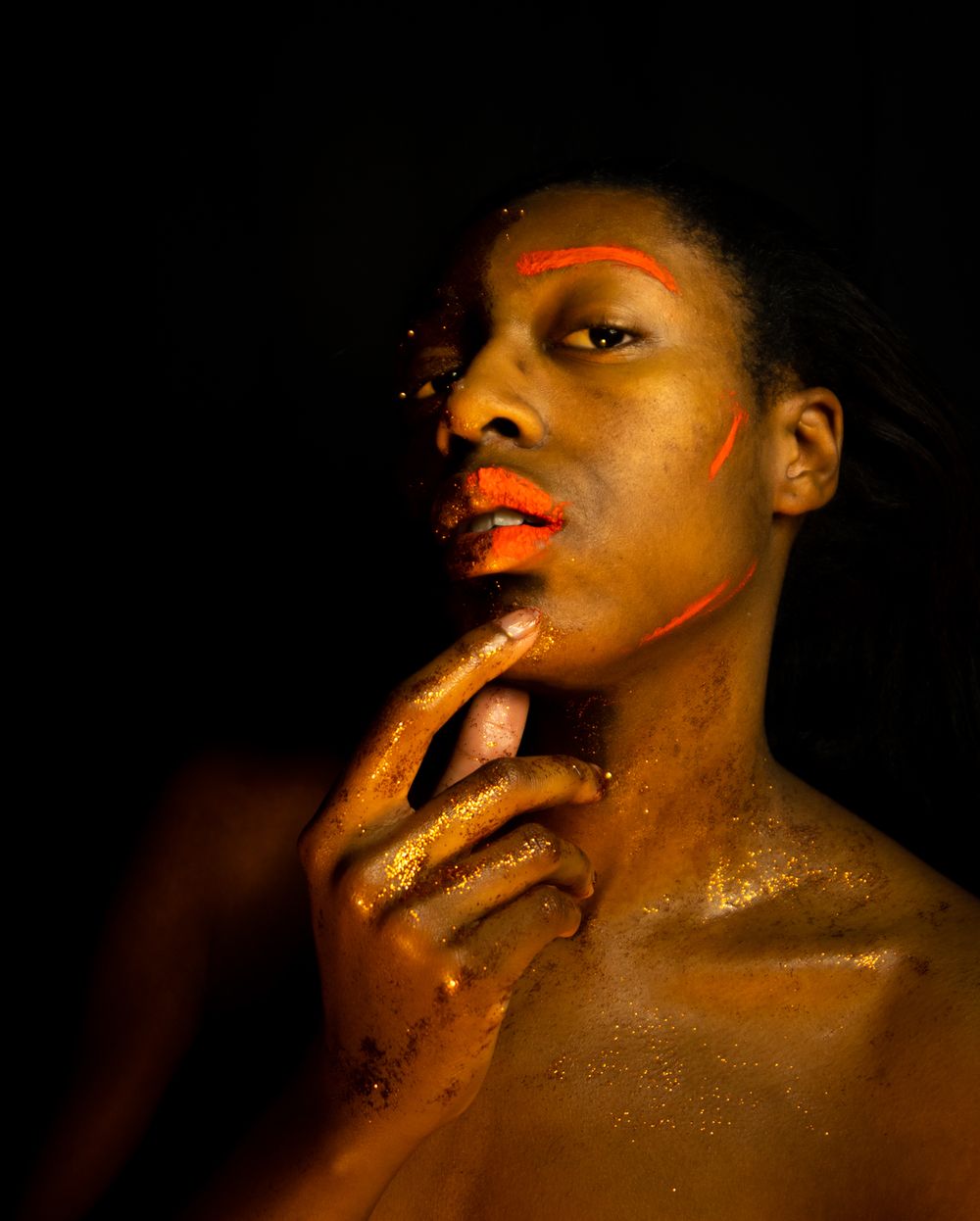 A person with orange paint on their face and gold glitter on their chest with their hand on their chin.