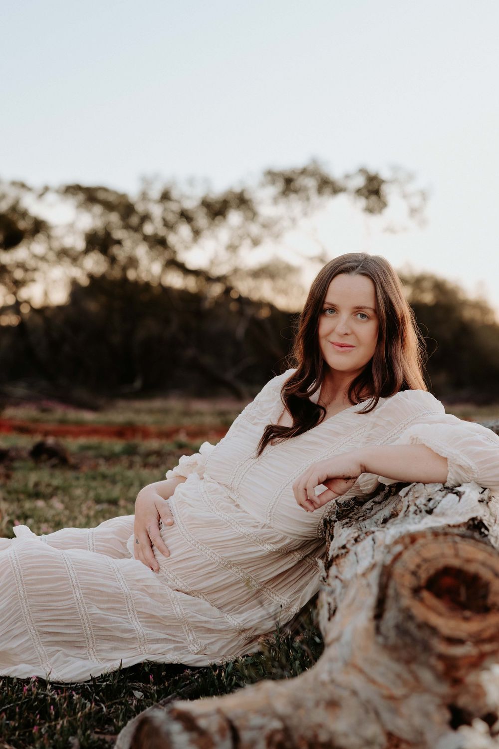 Woman in white dress reclining on fallen log in natural outdoor setting during golden hour.