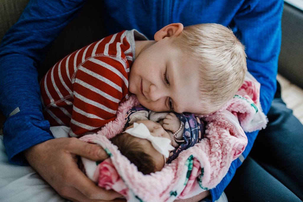 Young child in striped shirt cuddles with newborn baby wrapped in floral blanket.