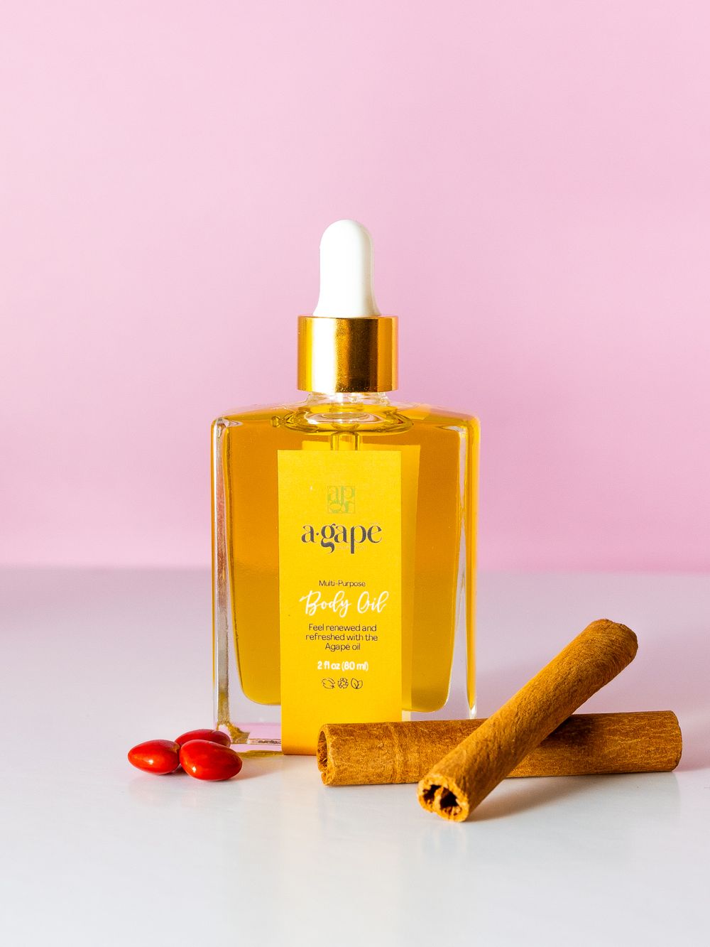 A bottle of golden Agape beauty oil with cinnamon sticks and red beads against a pink background.